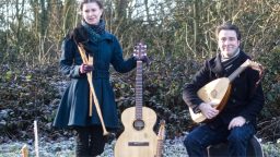 Musicians Sophie Matthews standing with a flute and guitar and Chris Green holding a mandolin in a cold wintery garden