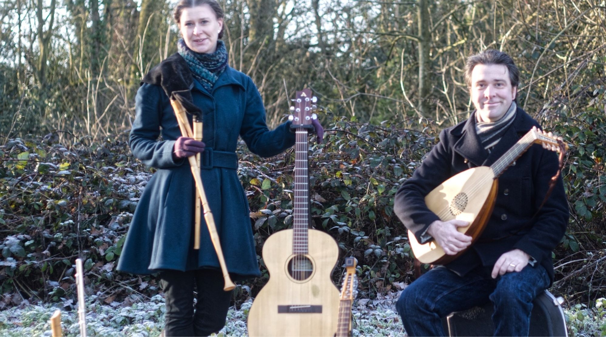 Musicians Sophie Matthews standing with a flute and guitar and Chris Green holding a mandolin in a cold wintery garden