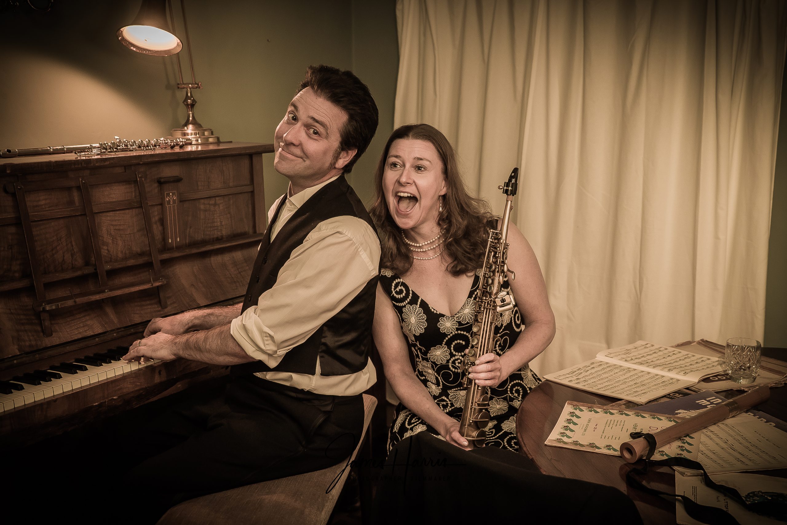 Musician Chris Green sat at a piano alongside Sophie Matthews who is holding a woodwind instrument. They are about to perform a song in a room with subtle lighting